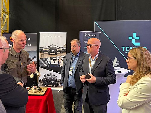 Ein Mensch in militärischer Uniform Unterhält sich mit vier weiteren Menschen auf einem Messestand.